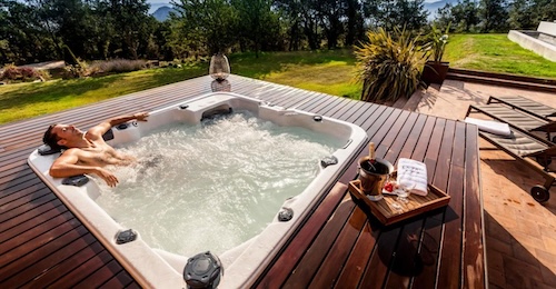 Un hombre relajado en el jacuzzi exterior en medio del campo sobre una plataforma de madera y con una botella y dos copas. 