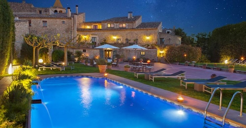 Piscina exterior con tubonas y el hotel Racó de Madremanya al fondo con paredes de piedra y estilo rústico iluminado.