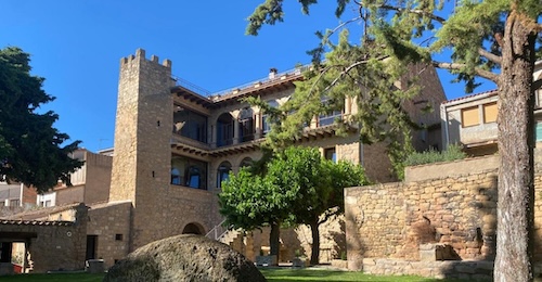 Foto del jardin exterior con una gran piedra, árboles y el hotel Casa Llobera 1792 con estilo rústico y de piedra al fondo.