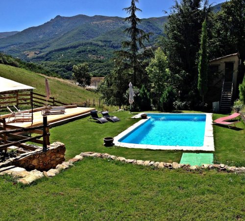 Foto de la piscina en medio del cesped junto a la casa con una terraza de madera con sombrilla y tumbonas con el bosque y las montañas al fondo