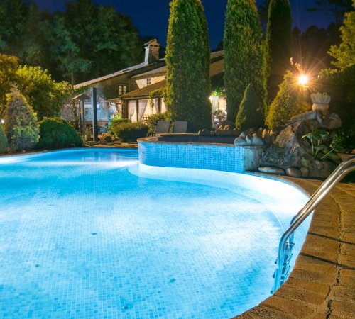 Foto de la piscina iluminada donde se ve la casa de estilo rústico y con árboles al fondo