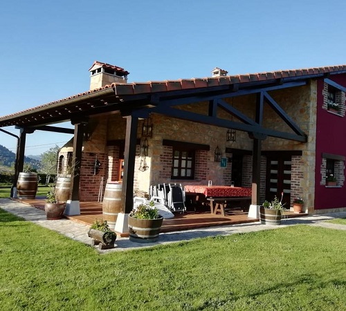 Vista de la casa rural La Milonga desde el exterior donde se ve el césped, las montañas al fondo y fachada de la casa en piedra