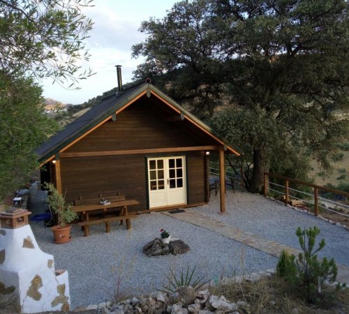 Foto de la Cabaña La Encina donde vemos las mesas del exterior, la zona de barbacoa, los árboles de alrededor y la cabaña construida en madera