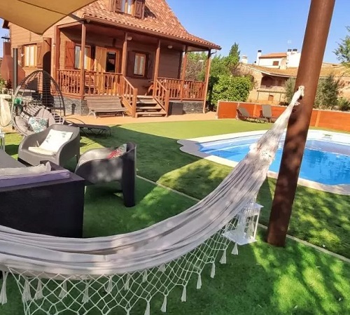 Vista del exterior de Casa Rural Casita de Madera donde se ve el exterior en madera, la piscina, el cesped y el mobiliario de la terraza donde comer.