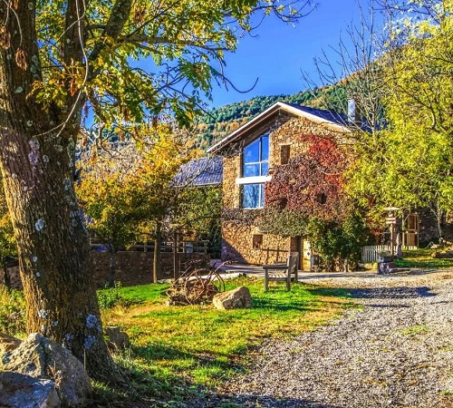 Vista desde el exterior con los árboles del jardín y la Casa Rural Cal Sodhi al fondo con exterior de piedra.
