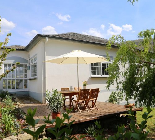 Exterior de Casa Dodo donde tienes una terraza con suelo de madera en el exterior con mesas y silla bajo una sombrilla rodeado de jardines y árboles
