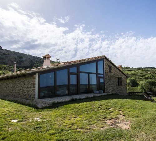 Vista desde el exterior de Masí el Cabrero, una casa rural en medio del campo con prados y colinas al fondo.