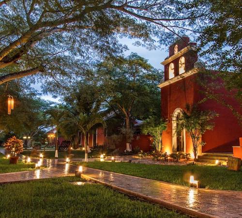 Vista de los jardines exteriores con las luces en el borde del camino y el hotel Hacienda Santa Cruz Merida al fondo con la fachada de color rojo
