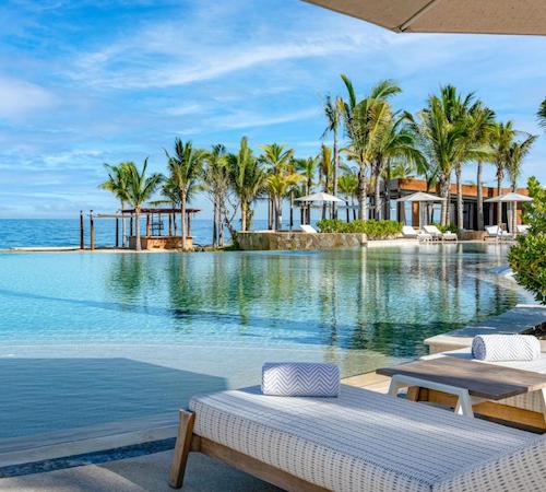 Dos hamacas para tomar el sol debajo de una sombrilla frente a una piscina con borde infinito con el mar de fondo en el Hotel Conrad Punta de Mita