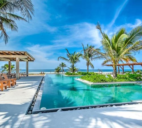Piscinas unidas con palmeras en su interior junto a unas sillas para tomar el sol y con vistas al mar en el hotel Marea Beachfront Villas