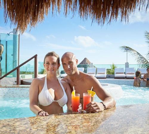 Pareja dentro de la piscina tomando una copa mientras vemos otras dos parejas al fondo desde donde se puede ver el mar.