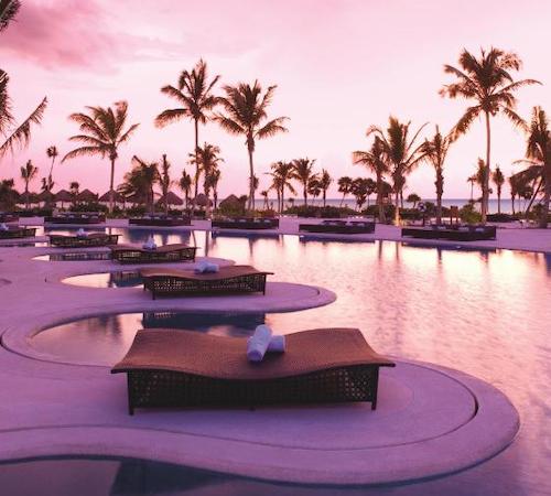 Piscina con muchas curvas y camas especiales para contemplar la puesta de sol sobre el mar y con las palmeras al fondo.