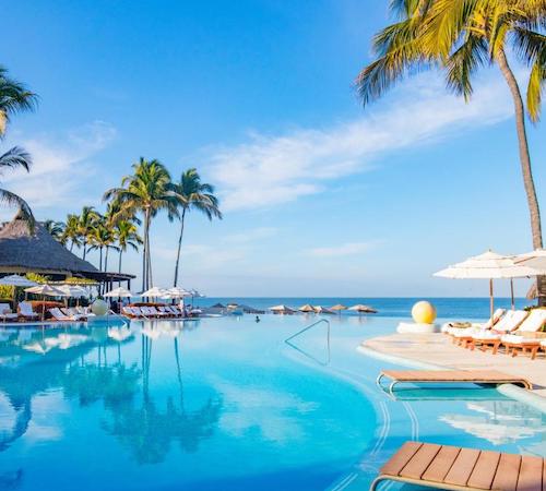 Vista de la piscina gigante rodeada de palmeras y tumbonas para tomar el sol y desde la que se puede ver el mar al fondo desde la piscina.