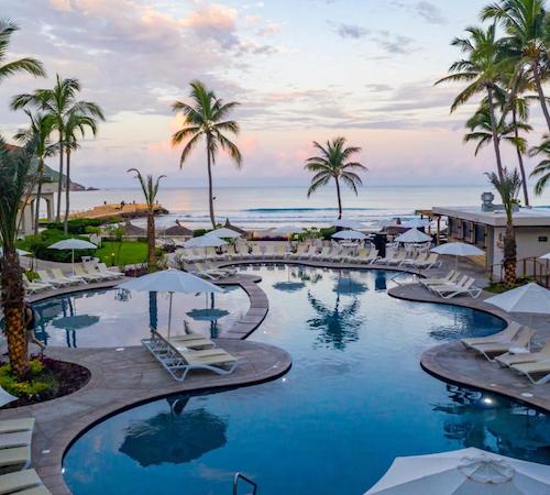 Piscina con vistas al mar con curvas y puentes para comunicar las orillas y rodeada de tumbonas para tomar el sol rodeado de palmeras