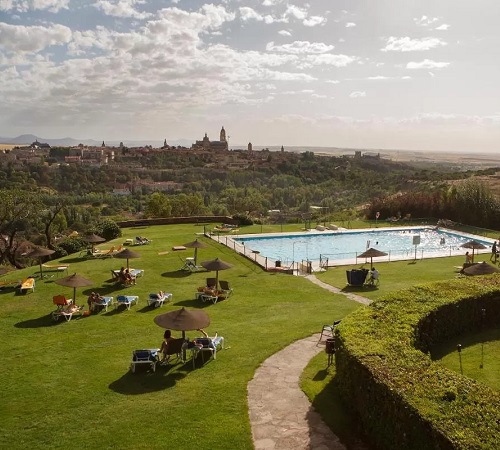 Jardines del Parador de Segovia con mesas en el césped, sombrillas y una piscina con vistas al ciudad