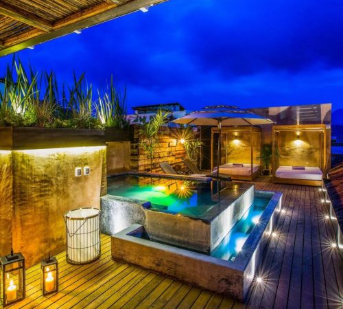 Vista de la terraza de suelo de madera donde se puede ver la piscina iluminada de noche con las camas balinesas al fondo en Múbú Hotel Boutique