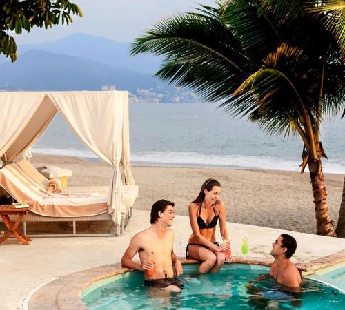 Dos chicos y una chica tomando una copa en el jacuzzi frente a una cama balinesa junto a la playa en el hotel Casa Velas – Adults only