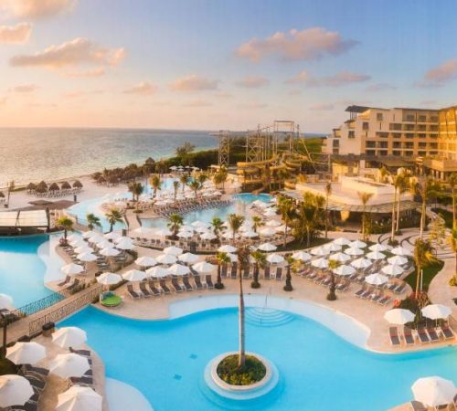 Foto de las dos piscinas con el puerte sobre ellas rodeado de las habitaciones del Hotel Dreams Natura Resort & Spa - All Inclusive y donde vemos la playa al fondo.