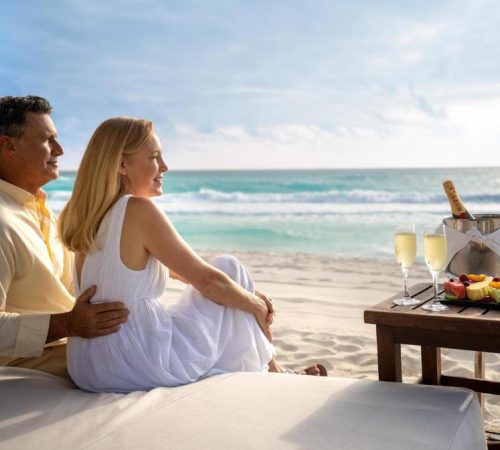 Foto de una parejas sentado mirando el mar con una botella de Champagne y una bandeja de fruta