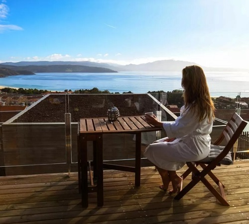 Foto de una mujer desayunando en la terraza de madera con vistas al mar en Cabañas Fisterra